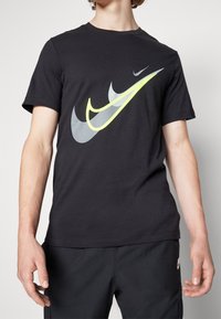 Černé bavlněné tričko s velkým reflexním logem Nike ve šedé a jasně žluté barvě. Krátké rukávy a kulatý výstřih.