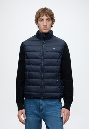 Junger Mann trägt eine marineblaue gesteppte Weste über einem schwarzen Langarmpullover und blauen Jeans, stehend vor einem einfarbigen weißen Hintergrund.