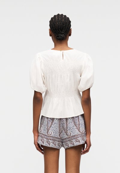 Femme aux cheveux tressés en chignon bas, portant un chemisier blanc à manches bouffantes et un short à motifs, debout de dos à la caméra.