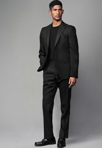 Schwarzer Anzug für Herren aus strukturiertem Stoff, kombiniert mit einem schwarzen Rundhalsausschnitt-Shirt. Merkmale: Slim Fit, aufgeschlagene Revers und glänzende schwarze Mokassins.