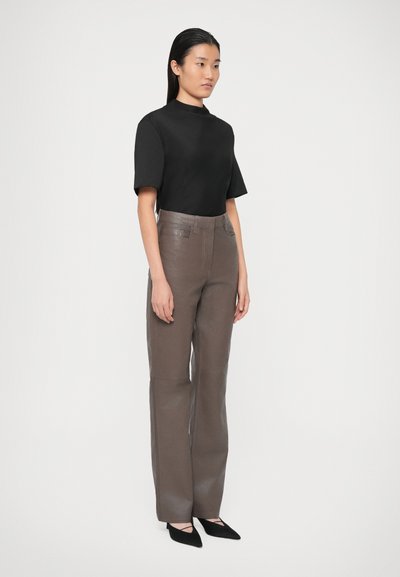 Vrouw staat en draagt een zwart short-sleeve top, taupe hoge leren broeken en zwarte puntige hakken met enkelbandjes.