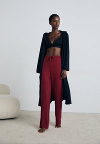 Svart lang cardigan over en svart bralette og løse røde bukser med snøring. Myk, bølgende stoff; uformell, avslappet passform.