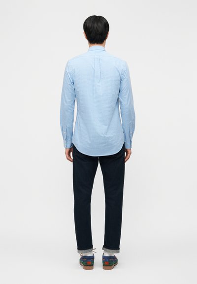 Man wearing long-sleeve blue checked shirt, dark jeans, gray socks, and colorful sneakers, standing facing away against white background.