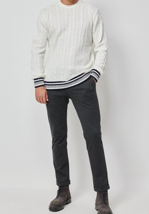 Weißer gestrickter Pullover mit Zopfmuster, ausgestattet mit navyfarbenen gestreiften Bündchen und Saum. Kombiniert mit dunkelgrauen Hosen und dunklen Stiefeln.