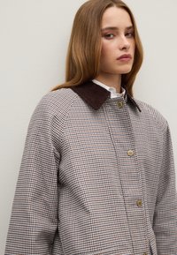 Bruine en witte houndstooth jas met een donkerbruine corduroy kraag, drukknoopsluiting en raglanmouwen. Zachte textuur en een ontspannen pasvorm.