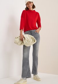 Haut rouge à manches longues avec un col haut et des détails de boutons, associé à un jean gris évasé et des baskets beiges, tenant un sac doux de couleur claire.
