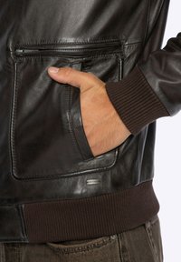 Bruine leren jas met een ritssluiting op de borstzak, geribbelde gebreide manchetten en een logotag. Het materiaal heeft een gladde, glanzende textuur.