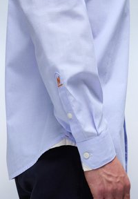 Lichtblauw langemouwen shirt van geweven katoen, voorzien van knopenmanchetten en een oranje "N" logo op de mouw, met een afgeronde zoom. 