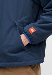Σκούρο μπλε μπουφάν με υφή, που διαθέτει ελ αστικά μανίκια και ένα εμφανές πορτοκαλί λογότυπο κοντά στον καρπό. Το χέρι βρίσκεται στην πλαϊνή τσέπη.