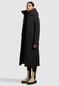 Lang sort dunjakke med høj krave, med lynlås og sidelommer, båret over sorte leggings, suppleret med beige ankelstøvler.