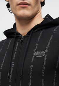 Sudadera negra con cremallera que presenta un patrón sutil de logotipo repetido. Tiene capucha con cordón y un parche circular de la marca en el pecho.