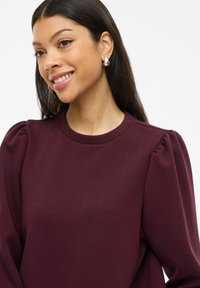 Bordeaux genser med puffermer og rund hals. Myk tekstur, tettsittende liv, og ribbede mansjetter. Sølvøredobb synlig.