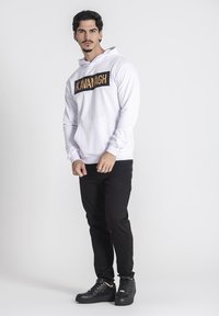 Biely mikina s logom "KAVANAGH" v čiernej farbe naprieč hrudníkom, predné klokanie vrecko, v kombinácii s čiernymi džínami a čiernymi teniskami.