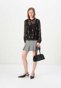 Donna che indossa una blusa nera trasparente a fiori e una gonna grigia a pieghe, tiene in mano una borsa nera ed indossa dei tacchi a slingback neri, mentre si trova su un pavimento bianco.