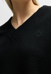 Schwarzer V-Ausschnitt-Pullover aus weichem Material mit geripptem Kragen und dezentem Logo-Detail auf der Brust, der ein klassisches Design präsentiert.