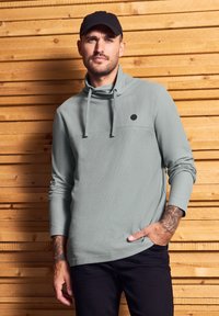 Světle šedá, texturovaná mikina s dlouhým rukávem a klokanní kapsou, zdobená logem, v kombinaci s černými kalhotami, umístěná na dřevěném pozadí.