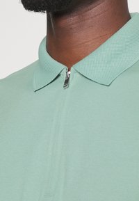 Polo shirt vert clair avec un col texturé et une fermeture éclair courte au niveau du cou. Tissu lisse avec un subtil éclat.
