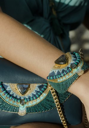 Bracelet et pochette assortie avec perles et pierres turquoise, or et bleu, porté au poignet sur un fond de tissu turquoise foncé.