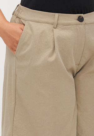 Close-up of person wearing beige pants with a button closure and hand resting in the front pocket.