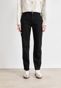 Mustad denim püksid sirge lõikega, millel on nööbisisu ja viis taskut, sobitatud heleda värvusega tennisekingadega.