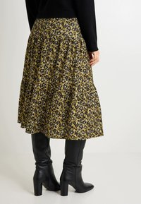 Falda floral en verde oliva con patrones en blanco y negro, con costuras en capas. Combinada con botas negras de tacón hasta la rodilla.