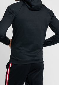 Felpa nera con cappuccio, design aderente, texture liscia, con maniche raglan. Abbinata a pantaloni neri con strisce laterali rosse.