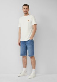 T-shirt blanche à manches courtes avec un petit logo de flamme orange, associée à un short en denim bleu à mi-cuisse et des baskets blanches. Le fond est neutre.