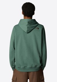 Grüner Kapuzenpullover mit glatter Textur, ausgestattet mit einem kleinen braunen Logo auf dem oberen Rücken und gerippten Bündchen am Saum.