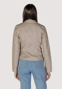 Giacca in ecopelle beige con un design aderente, spalline sulle spalle e cuciture posteriori. Abbinata a jeans azzurri a vita alta.