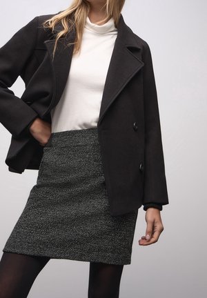 Frau trägt einen schwarzen zweireihigen Mantel, weißen Rollkragenpullover, schwarz-weiß gemusterten Rock und schwarze Strumpfhosen vor einfachem Hintergrund.