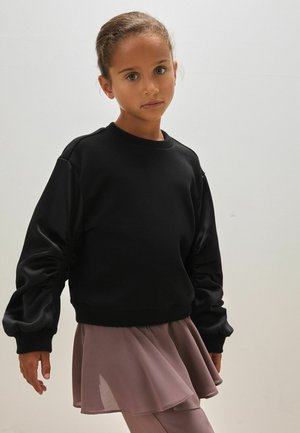 Młoda dziewczyna o brązowych włosach związanych z tyłu nosi czarną oversize bluzę i mauve spódnicę, stojąc na jasnym, jednolitym tle.