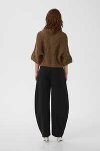 Γυναίκα με κόκκινα μαλλιά μήκους μέχρι τους ώμους φοράει καφέ χνουδωτό πουλόβερ με φαρδιές μανσέτες, μαύρο παντελόνι με φαρδιά μπατζάκια και λευκά παπούτσια με τακούνι, κοιτάζοντας πίσω.