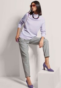 Lilafarbenes Strickpullover mit hohem Kragen, graue maßgeschneiderte Hose und lila Wildledersandalen. Große Perlenkette, übergroße Sonnenbrille.