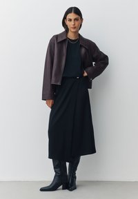 Giacca corta in pelle marrone, top nero, gonna nera, stivali neri al ginocchio con punte appuntite, dettaglio cintura con accenti dorati. Tessuti lisci.