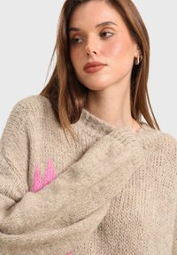 Pull en tricot beige à coupe ample, avec un grand motif rose à l'avant. Il a des poignets côtelés et un encolure large.