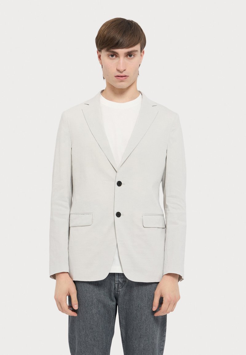 Helles graues Blazer aus glattem Stoff, mit zwei Fronttaschen, zwei schwarzen Knöpfen und taillierter Passform. Wird über einem weißen Hemd getragen.
