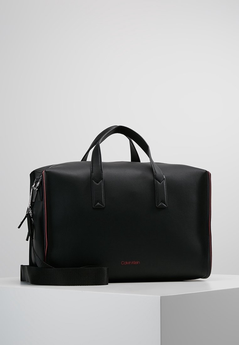 Sac de voyage en cuir noir avec deux poignées supérieures, une bandoulière amovible et des surpiqûres rouges. Présente un logo rouge sur le devant.