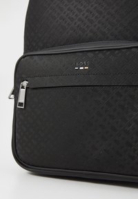 Sac à dos noir à motif avec une surface texturée, comportant une poche zippée et des accents en métal argenté. Logo affiché à l'avant.