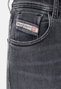 Vaqueros de mezclilla negra con un bolsillo delantero que lleva una etiqueta del logo de Diesel en rojo y blanco. La tela tiene una apariencia ligeramente texturada.