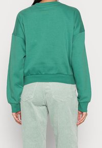 Felpa verde in cotone con spalle basse, scollatura tonda e polsini e orlo a costine. Indossata con pantaloni in corduroy verde chiaro.