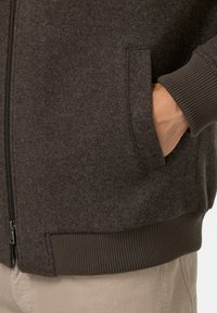 Tmavě hnědá zipová bunda z měkké a strukturované tkaniny, s bočními kapsami a vroubkovanými manžetami a lemem.