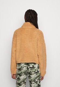 Veste en polaire beige, confortable et courte, avec un col haut, présentant une surface texturée et un détail de patch accentué au dos. Assortie avec un pantalon camouflage.