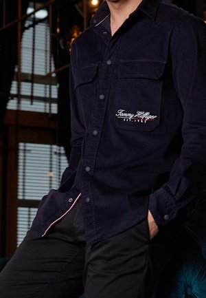 Personne portant une chemise bleu foncé boutonnée avec "Tommy Hilfiger EST. 1985" brodé sur la poche poitrine, mains dans les poches d'un pantalon noir.