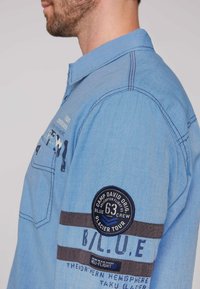 Camisa de manga larga de color azul claro con un cuello, que presenta rayas en azul oscuro y gris, parches bordados y una tela texturizada.
