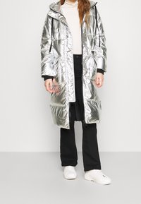 Manteau rembourré métallique argenté avec capuche, doté de poches latérales, d'une fermeture éclair à l'avant et de poignets noirs, porté sur un haut crème et un pantalon noir.