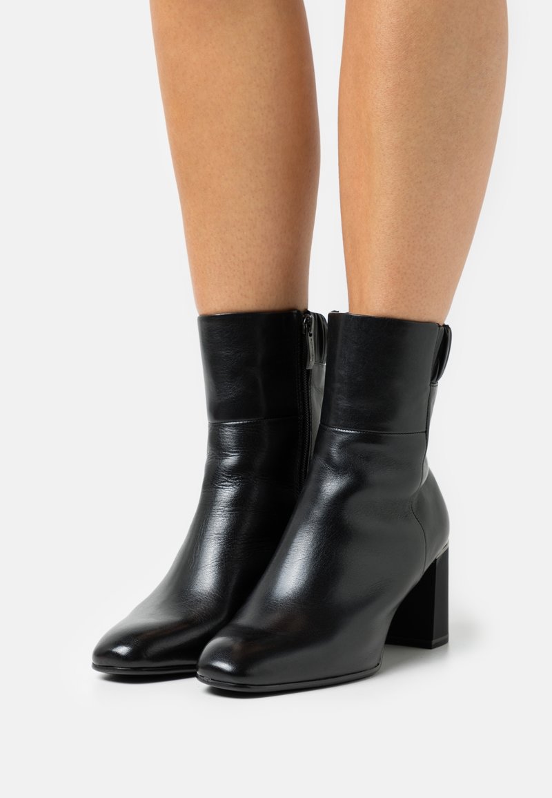 Bottines en cuir noir à talons carrés portées sur jambes nues sur un fond blanc uni.