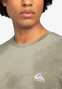 Camiseta de algodón verde con cuello redondo y un pequeño logo blanco de Quiksilver en el lado izquierdo del pecho. Textura suave, diseño casual.