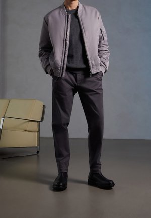 Mężczyzna ubrany w szarą kurtkę bomber, ciemny sweter, ciemne spodnie i czarne buty, stojący obok beżowego fotela w minimalistycznym otoczeniu.