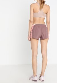 Sport-BH in Beige, kombiniert mit mauvefarbenen Laufshorts mit pinken Seitenpanelen. Modell trägt hellrosa Sportschuhe. Glattes, leichtes Material.