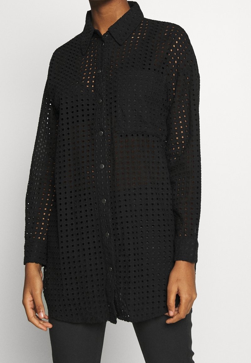 Chemise noire à manches longues avec boutons et motif de découpes carrées portée par une personne debout, les mains détendues le long du corps.